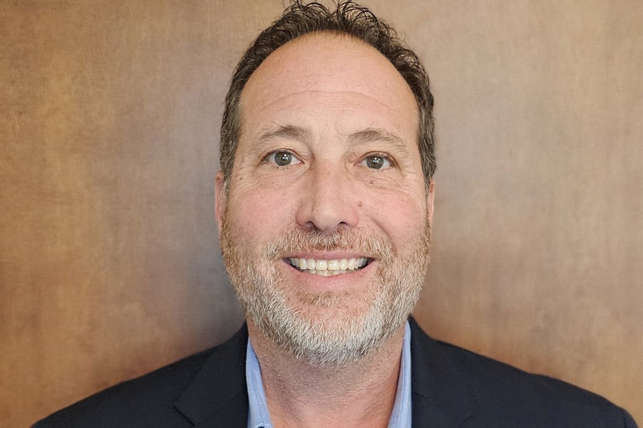 Headshot of Cary Goldberg, a middle-aged man in a dark suit and tie, smiling against a gray gradient background.