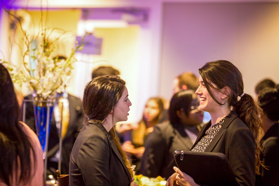 Two young professional women in conversation at a networking event, surrounded by other attendees in a warmly lit room.