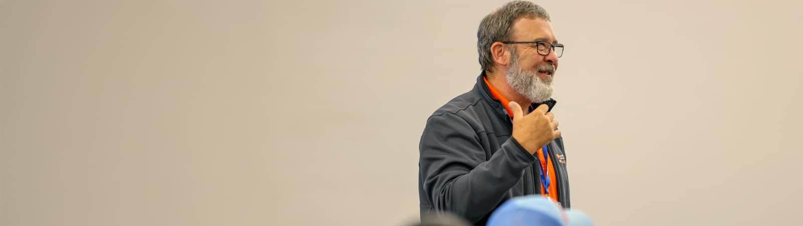 A man with glasses and a beard, wearing a dark jacket and orange lanyard, speaks animatedly to a group, gesturing with his right hand.