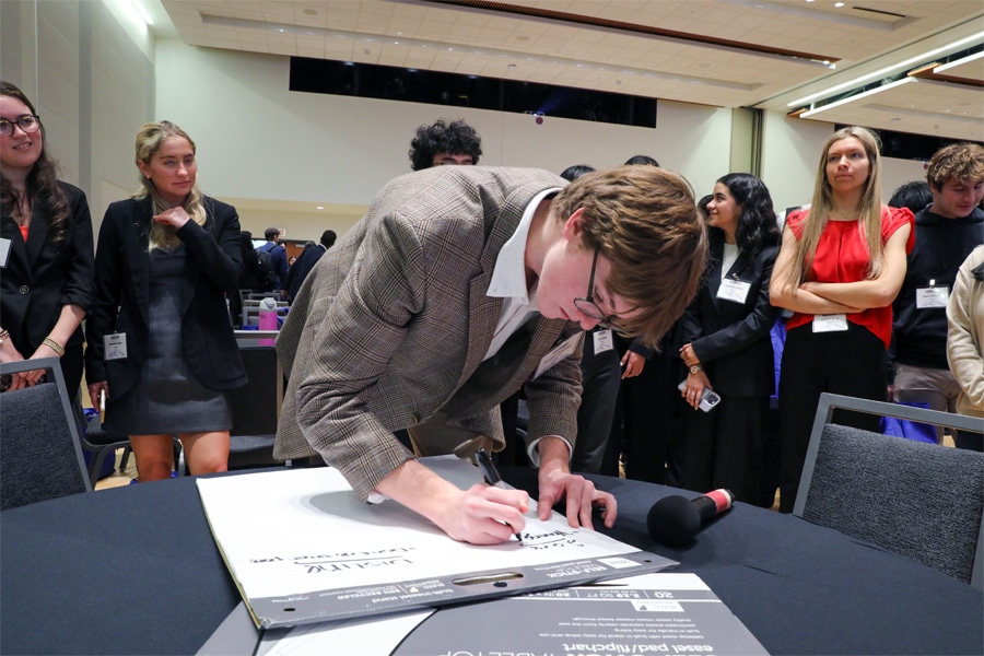 A young man in glasses and a plaid blazer leans over a table, signing a large poster. Behind him, a group of well-dressed students watches attentively in a conference room setting.