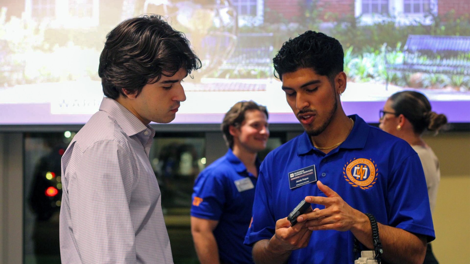 A Warrington Diplomat in a bright blue polo shirt helps a fellow student navigate information on a phone or device. They stand together in front of a screen showing a presentation, surrounded by other engaged students.