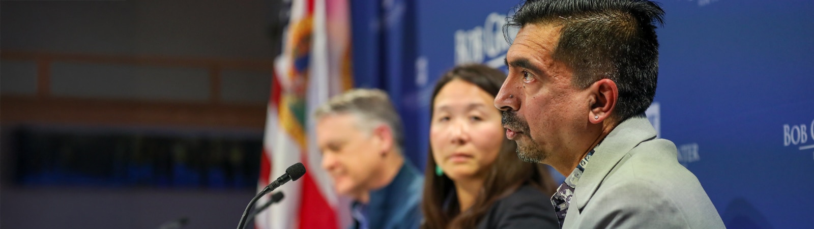 Three business professionals sit at a panel table with microphones, speaking and listening intently during a formal discussion. The American and Florida state flags are visible in the background.