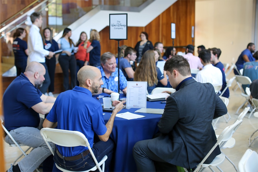Recruiters from The Walt Disney Company engage in conversation with graduate students seated at tables in a large networking event space, with other attendees mingling in the background.