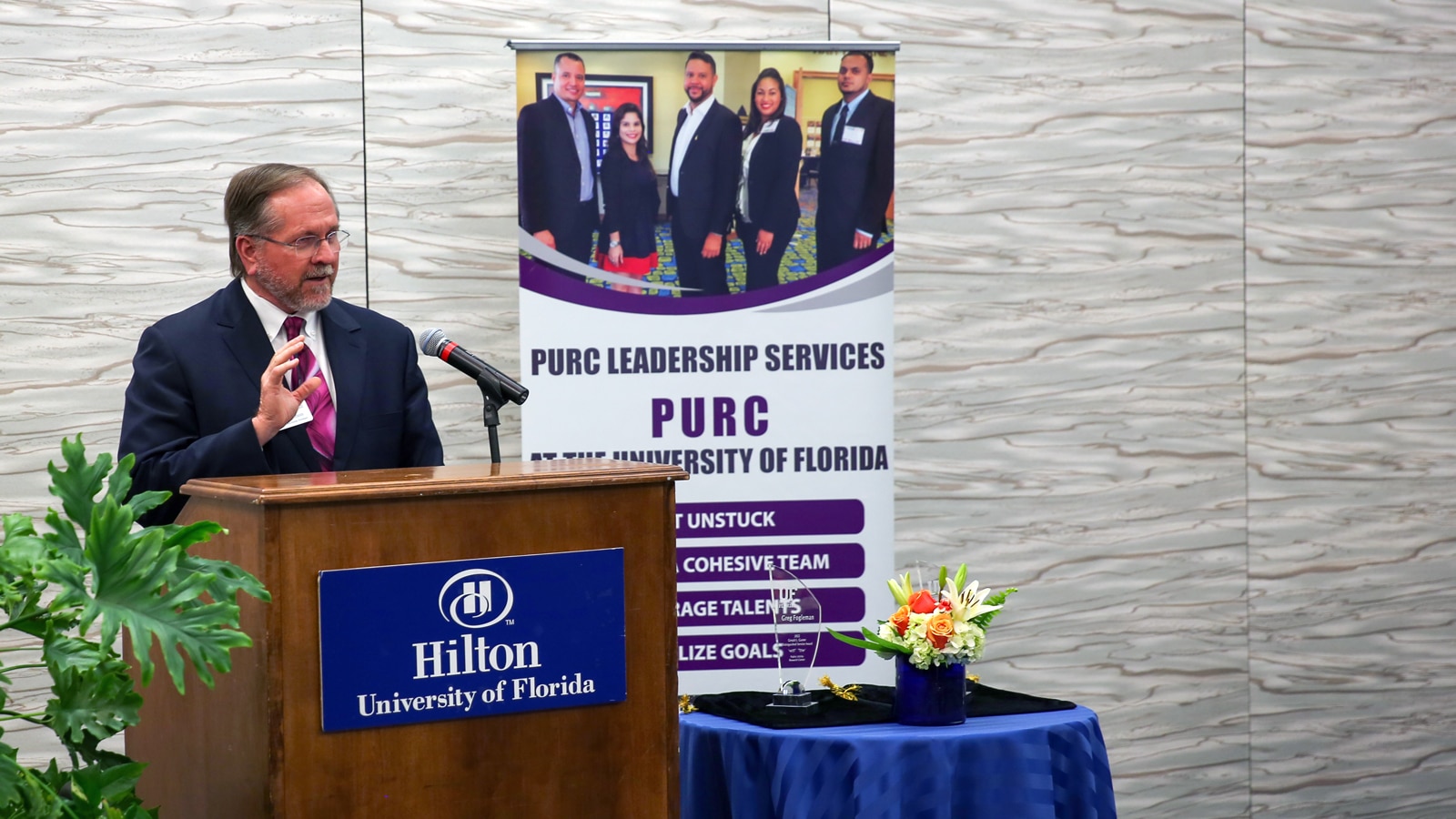 A speaker in a suit addresses attendees from a podium at the PURC Annual Conference, with a PURC Leadership Services banner and awards table in the background.