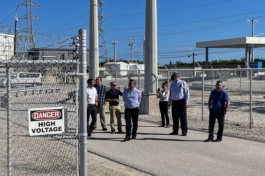A group of professionals tours an electrical utility substation, standing outside a high-voltage fence in bright, sunny weather.