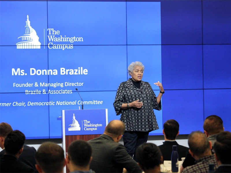 Donna Brazile, Founder and Managing Director of Brazile & Associates, speaks to a group of students at The Washington Campus, standing in front of a large blue screen displaying her name and credentials.