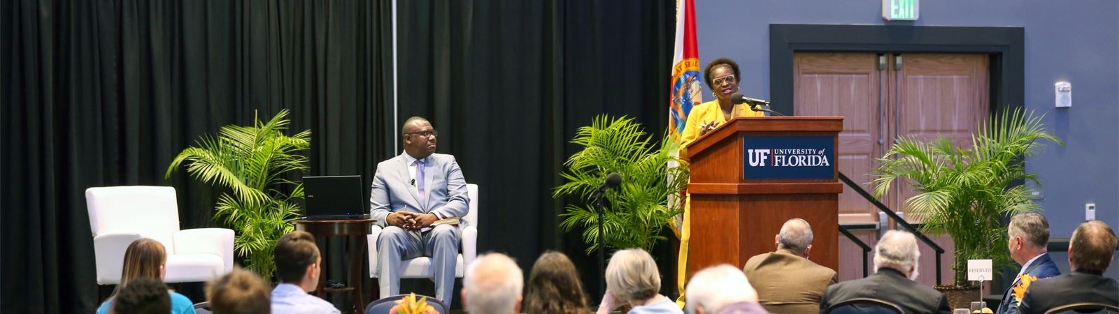 A speaker at a podium with a University of Florida sign addresses an audience in a conference setting, while a seated panelist listens attentively.