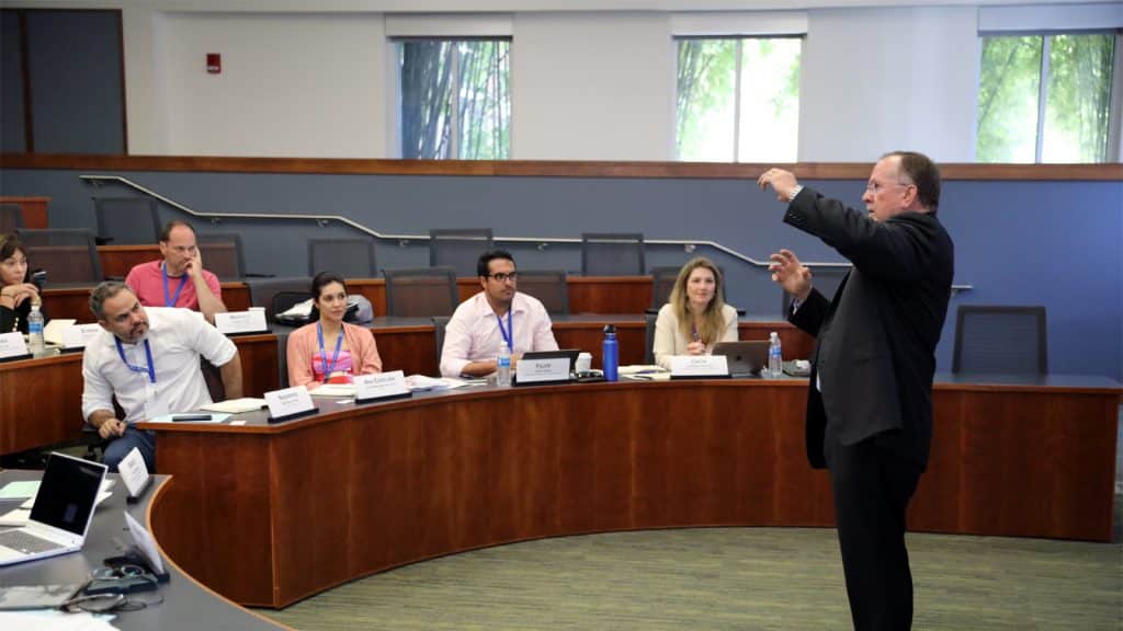 Mark Jamison stands at the front of a semicircular classroom, gesturing with his hands while addressing a group of attentive students seated behind curved desks.