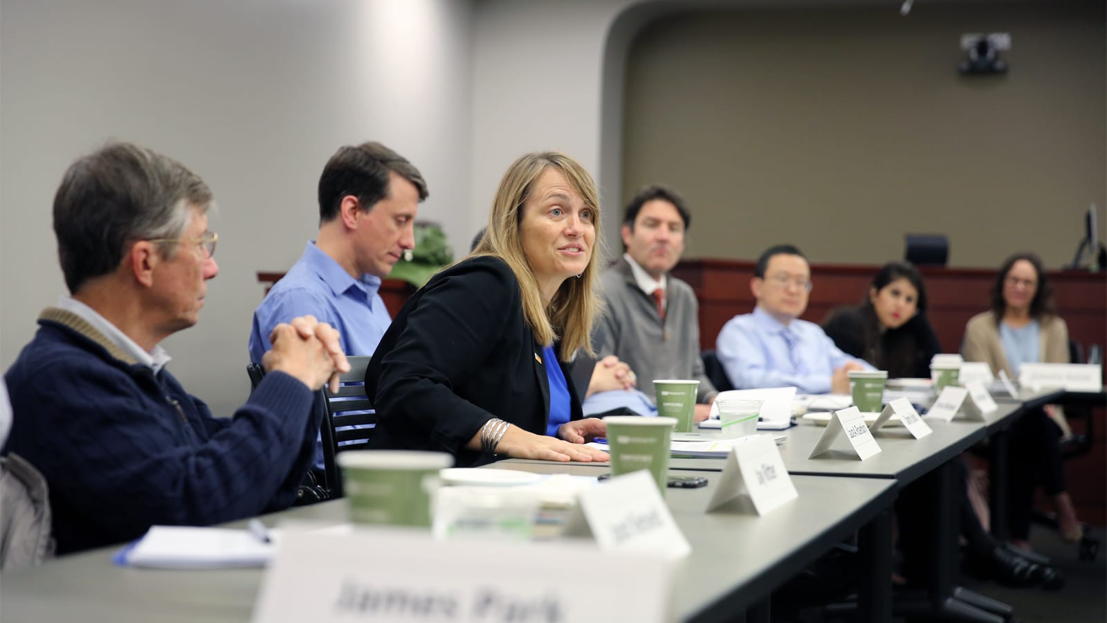 A woman in a black blazer speaks while seated at a conference table with several other participants in a meeting room.