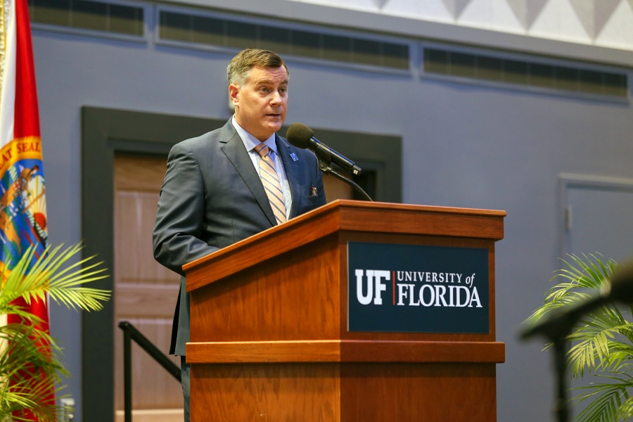 A man in a suit stands behind a wooden podium with the University of Florida seal, delivering a speech at an event.