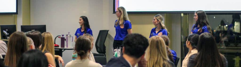 Several student ambassadors in blue Warrington Diplomats shirts stand at the front of a classroom, speaking to a seated audience of students.