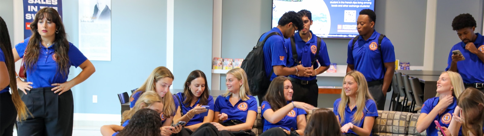 A diverse group of Warrington Diplomats in matching blue polo shirts chat, check their phones, and interact with one another in a modern building lobby, showcasing camaraderie and leadership.