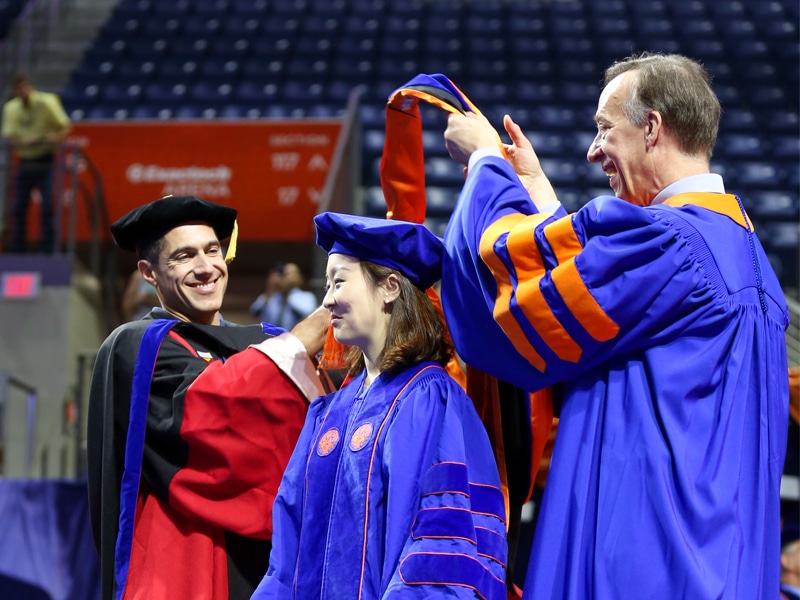 A doctoral graduate in a blue robe is hooded by two faculty members in academic regalia during the University of Florida doctoral ceremony.