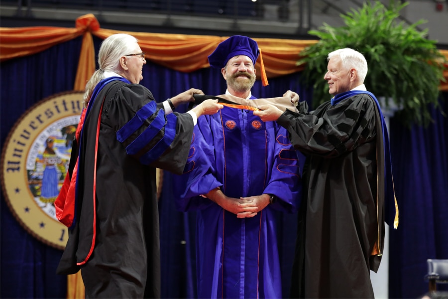 Two faculty members in academic regalia place a doctoral hood on a graduate dressed in blue doctoral robes and cap, standing on stage in front of the University seal.