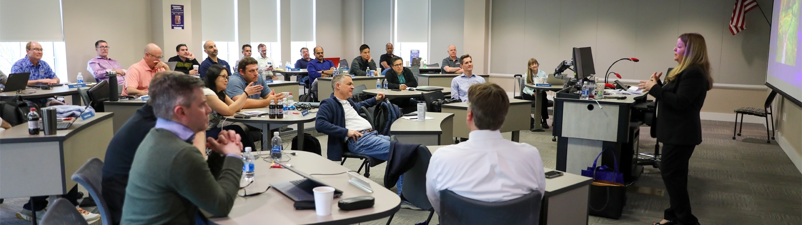 A classroom of adult students attentively listens to a female instructor presenting at the front of the room at the Warrington College of Business.