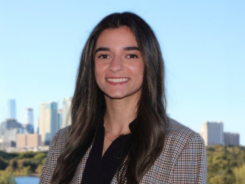 A young woman with long dark hair smiles confidently in a checked blazer with a black blouse underneath, with a city skyline in the background.