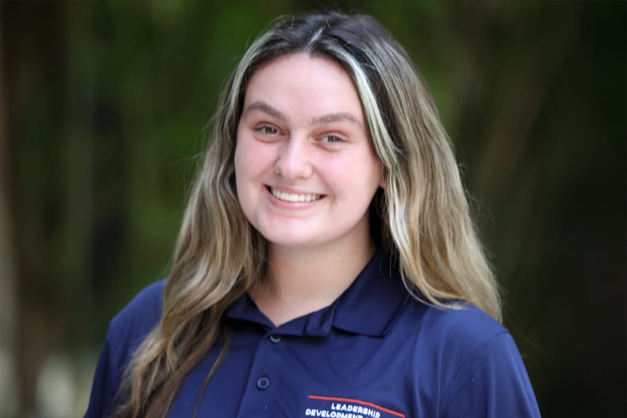 A young woman with long, wavy hair smiles confidently at the camera, wearing a navy blue Leadership Development Program polo shirt, with a blurred outdoor background.
