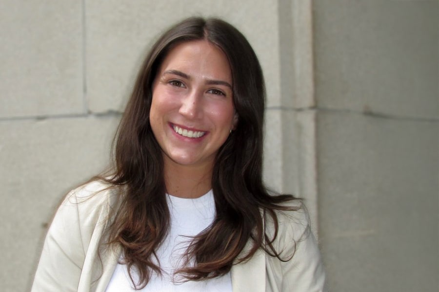 A young woman with long, dark brown hair smiles warmly at the camera, wearing a light-colored blazer over a white top, standing in front of a stone wall.