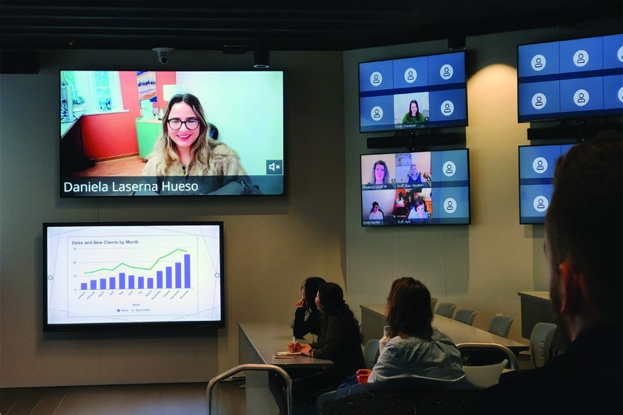 Students sit in a classroom watching multiple large screens displaying a video call with a woman named Daniela Laserna Hueso, a chart titled "Sales and New Clients by Month," and several smaller video call participant windows.