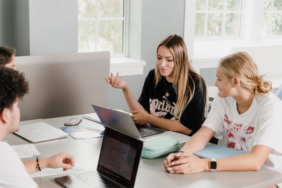 A group of students sitting around a table, engaged in discussion with laptops open and papers spread out.