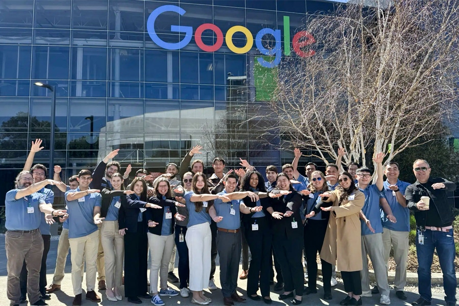 A large group of students and faculty stand in front of the iconic Google building in Silicon Valley, arms outstretched in a friendly pose. The bright Google logo is clearly visible above the group.