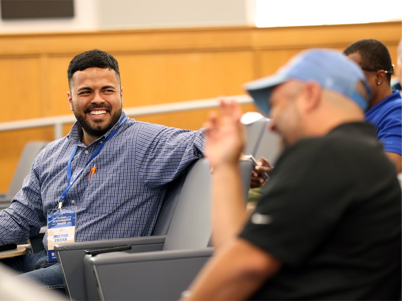 A smiling man wearing a blue checkered shirt and a lanyard with a name badge talks with another man in a baseball cap seated beside him in a classroom or auditorium setting.