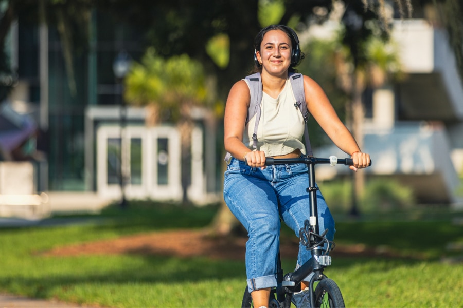 A smiling student wearing headphones and a backpack rides a bicycle across the University of Florida campus on a sunny day.