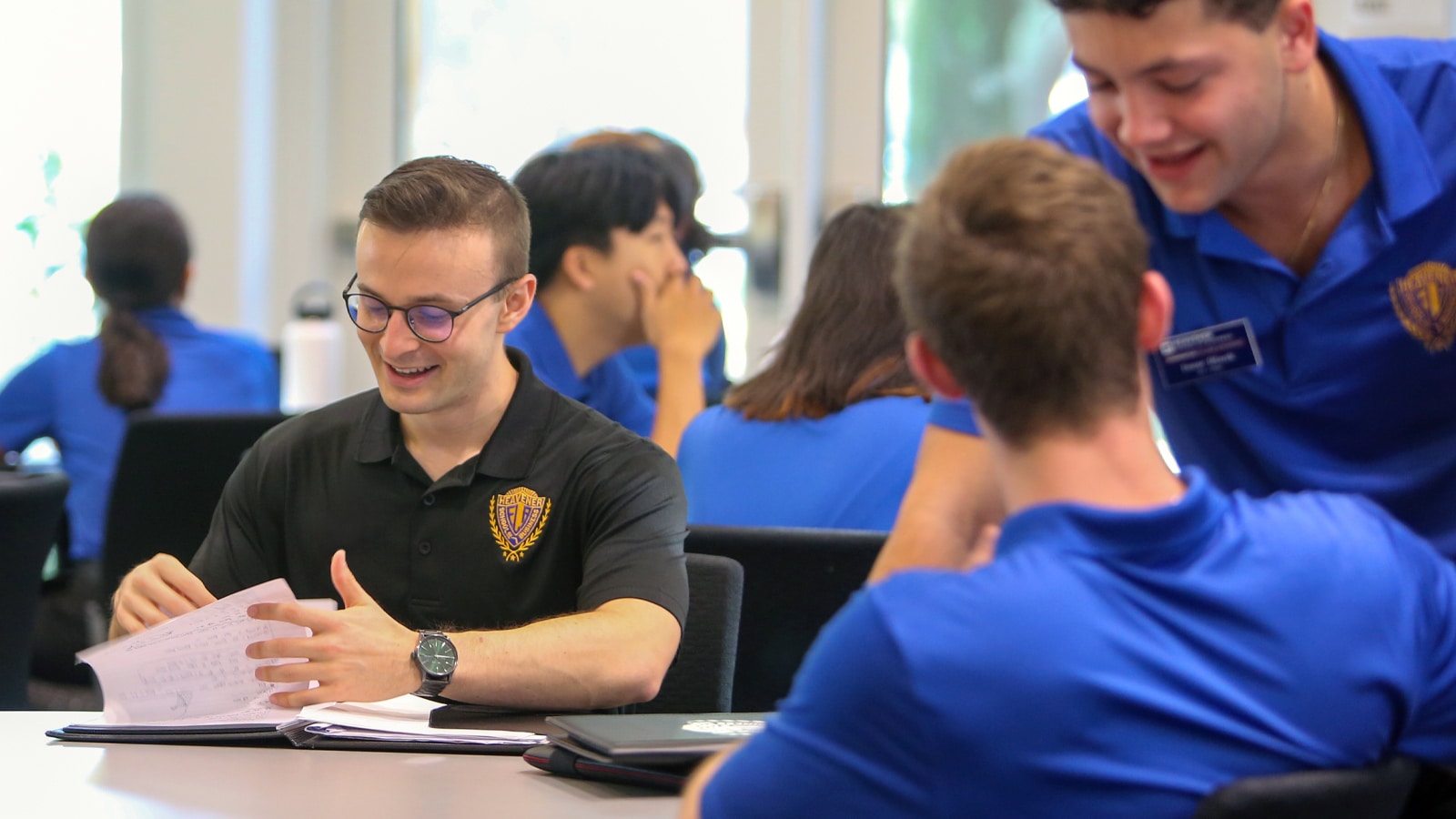A group of college students in a bright room. One student wearing glasses and a black shirt with the Heavener logo smiles while flipping through papers. Another student in a blue shirt leans forward talking with two other seated students.