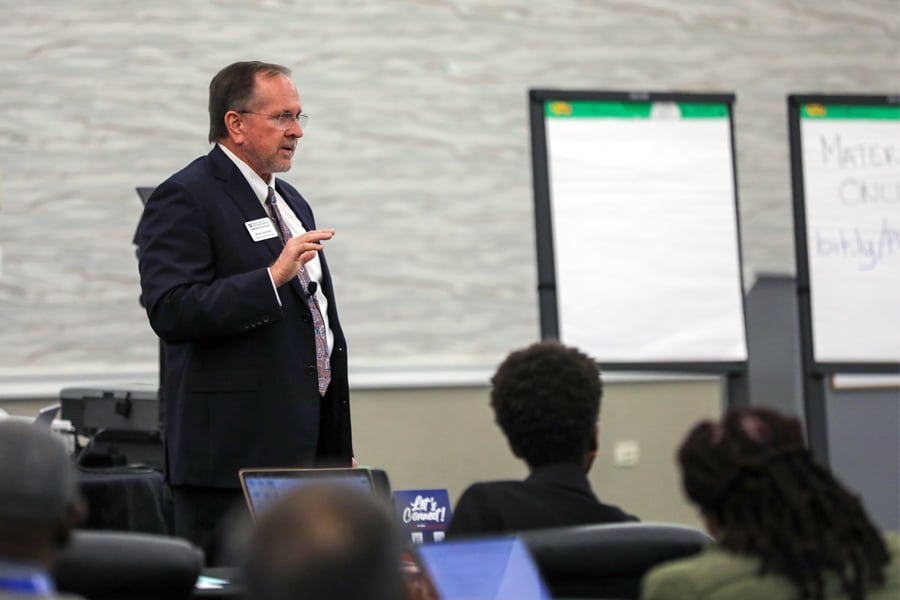 Mark Jamison, wearing a suit and tie, speaks at a workshop. He gestures with his hand as he addresses an audience seated before him.