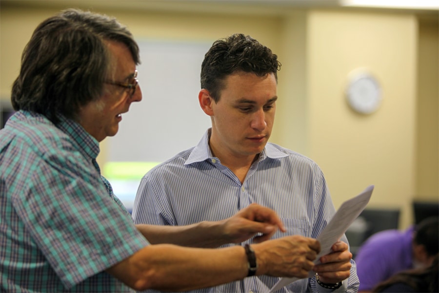 A faculty member in a checkered shirt points to a document while discussing it with a young man in a striped shirt in a classroom setting.