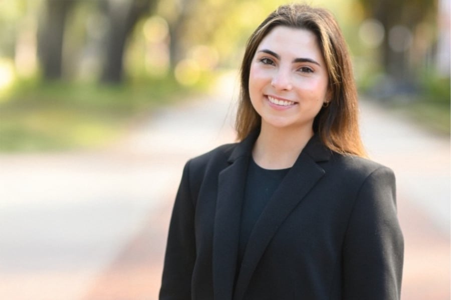 A young woman with straight brown hair smiles confidently at the camera, dressed in a black blazer, standing outdoors on a sunlit path with trees in the background.