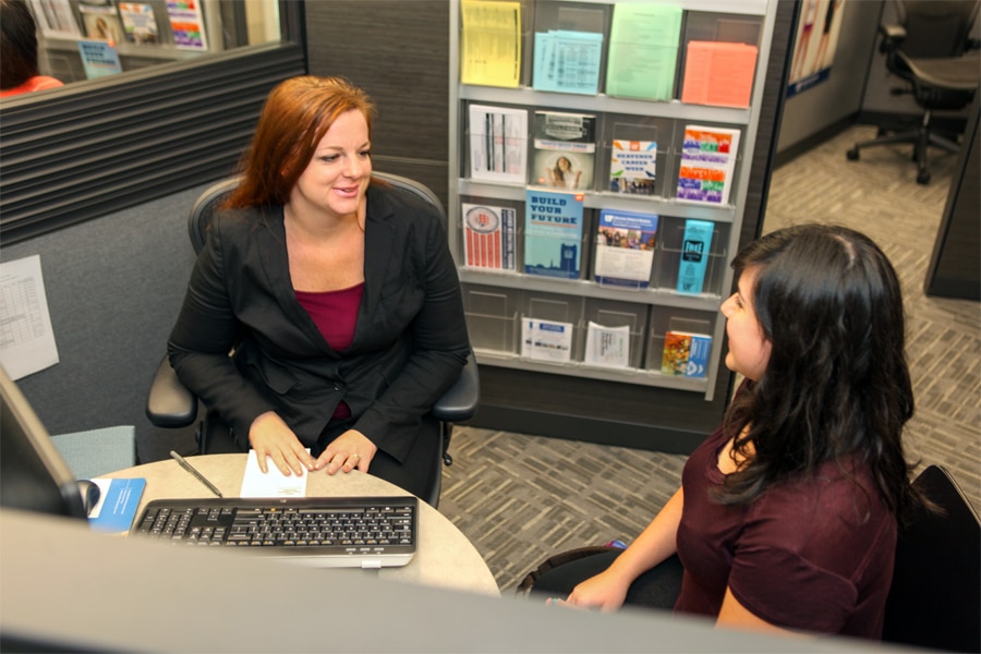 An academic advisor and a student sit across a desk in an advising office, reviewing degree requirements on a computer terminal.