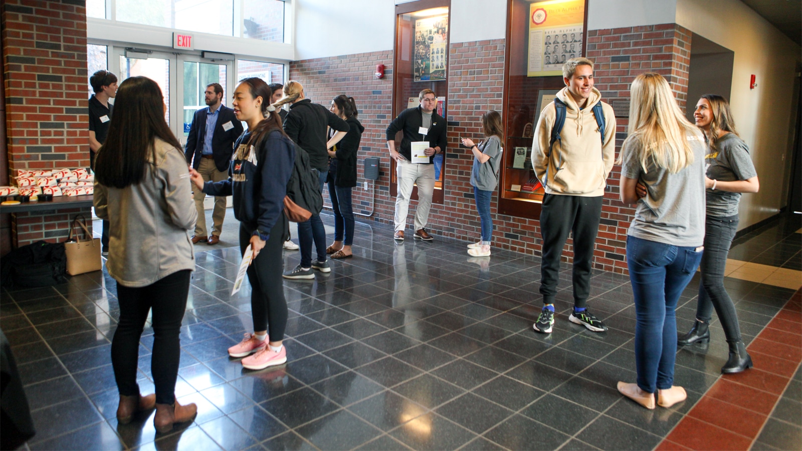 Groups of students and professionals engaged in conversation in a modern academic building lobby with brick walls and display cases.