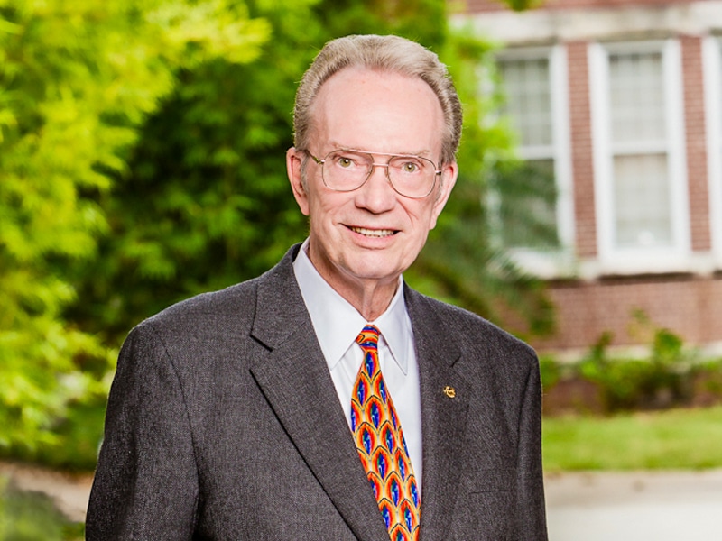 Portrait of Fred Fisher, an elderly man with glasses, wearing a gray suit and colorful tie, smiling in front of a leafy background and brick building.