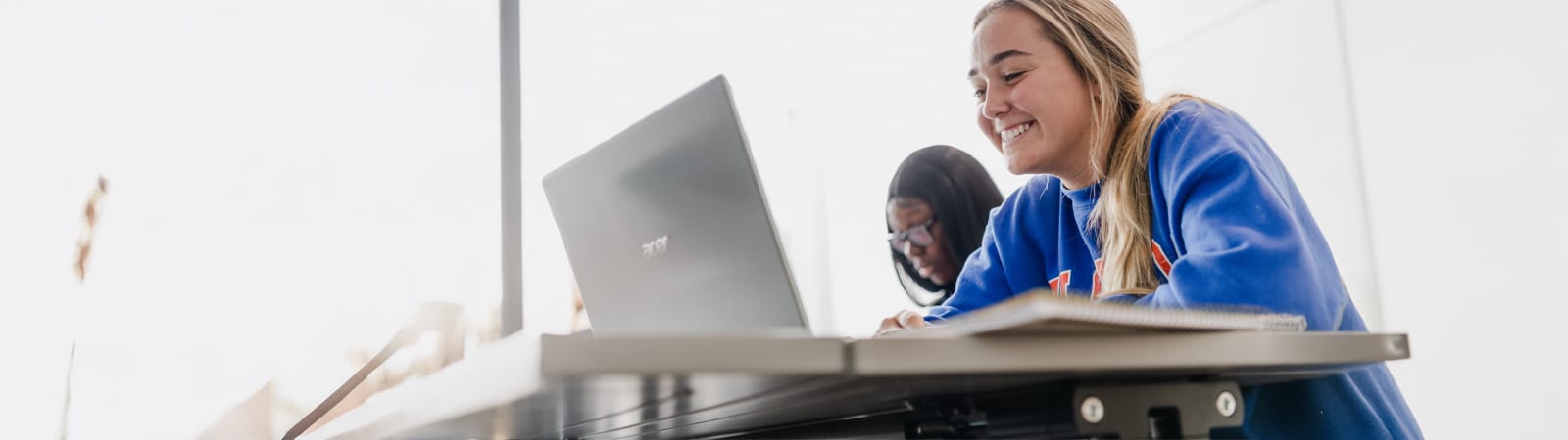 A student wearing a blue sweatshirt smiles while using a laptop, with another student working in the background.