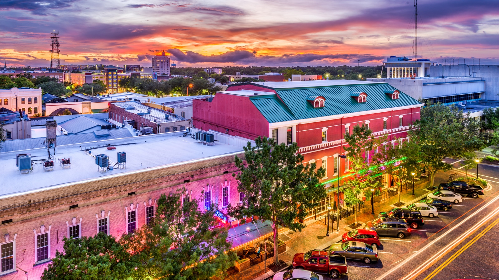 A colorful sunset over downtown Gainesville, Florida, with historic brick buildings, glowing neon lights, and tree-lined streets filled with parked cars.