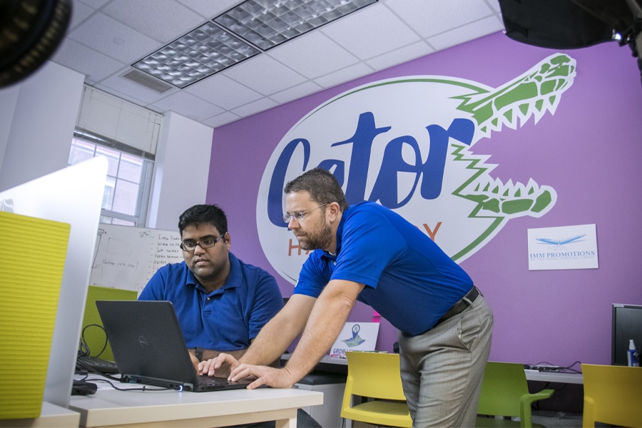 Two men collaborate at a laptop in a colorful office, with a large Gator Hatchery logo on the wall behind them.