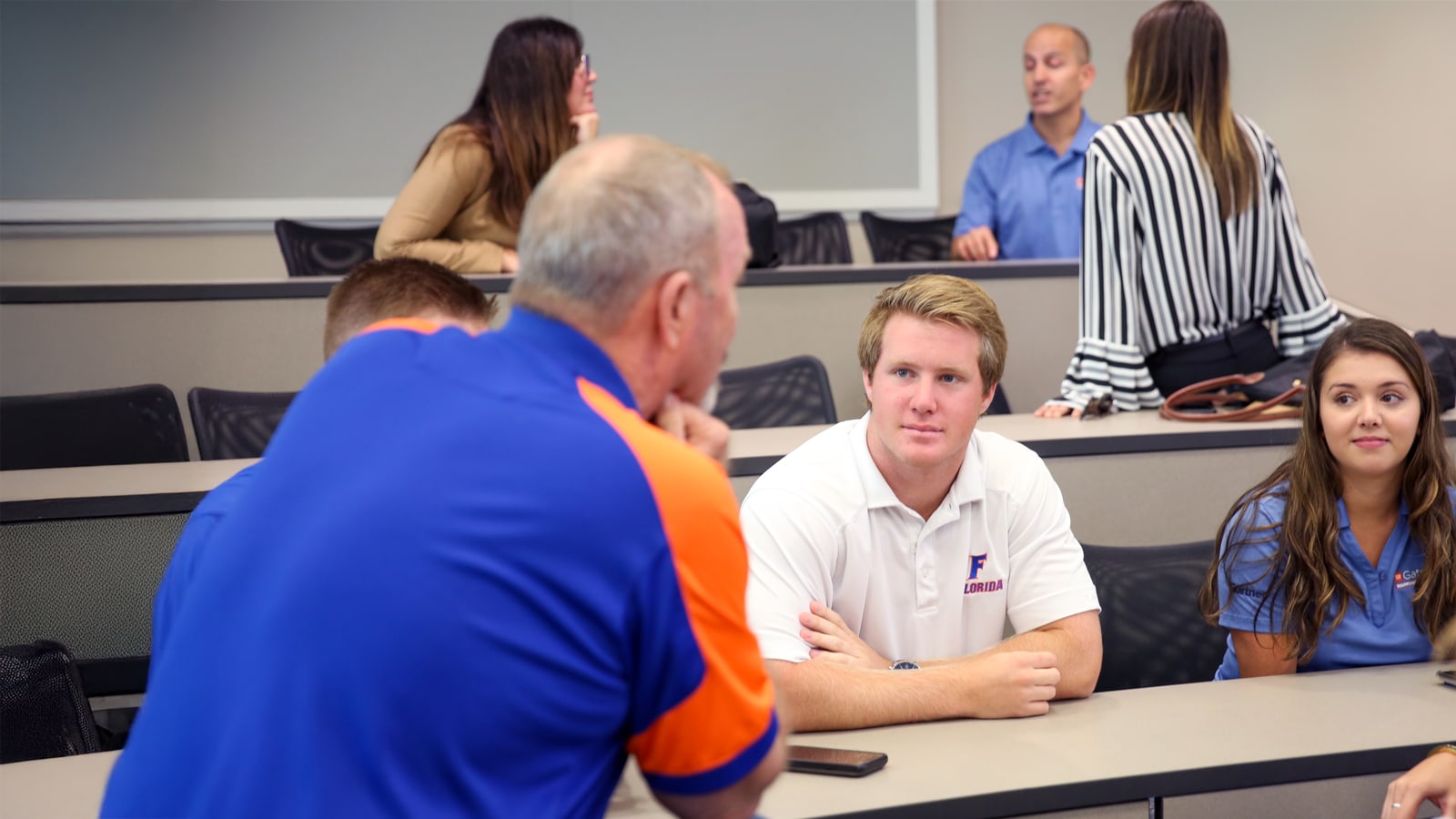 A student in a white Florida polo listens intently to a mentor in a blue and orange shirt during a classroom discussion, with other students and faculty interacting in the background.