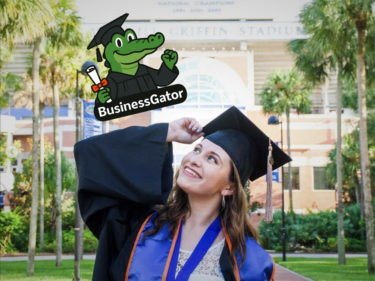 A smiling graduate in cap and gown looks up at a floating animated “BusinessGator” sticker showing the UF alligator in graduation regalia holding a diploma.