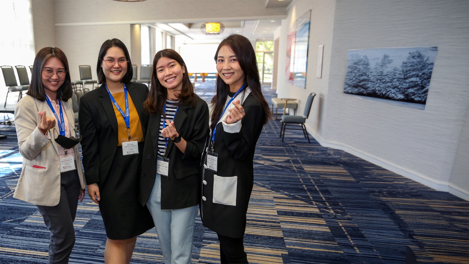 A group of four young women, dressed in business attire with conference name tags, smile and pose together at a professional event.