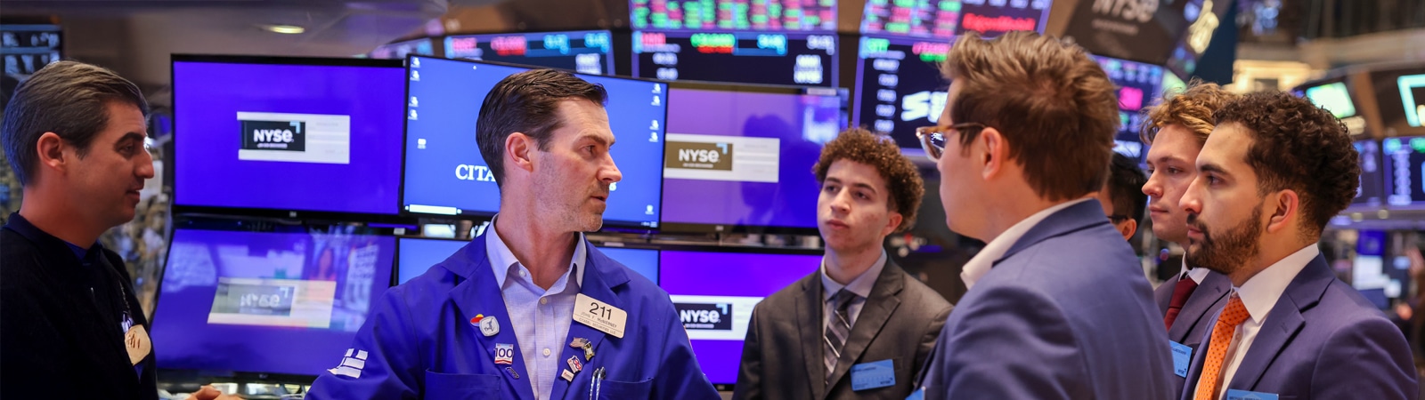 Graduate business students engage with a stock trader on the floor of the New York Stock Exchange, surrounded by monitors displaying stock information.