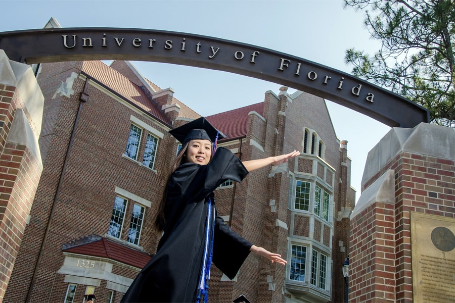 A University of Florida graduate in cap and gown poses under the iconic University of Florida archway, smiling and gesturing proudly.