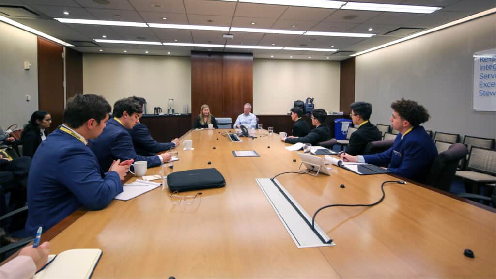 Graduate business students participate in a roundtable meeting, engaging in discussion with two professionals at the head of a large conference table.