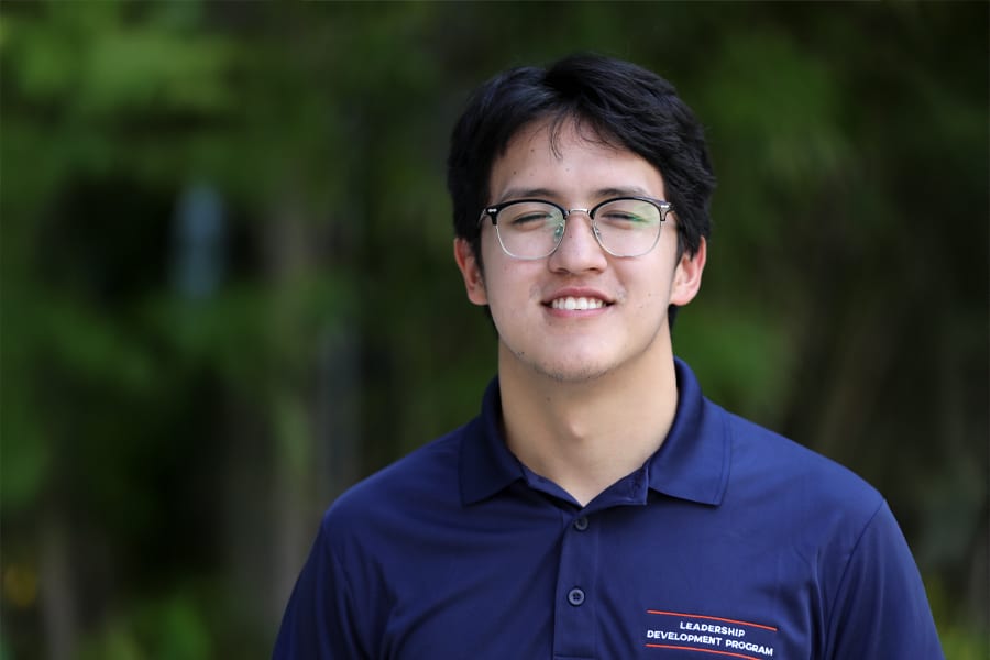 A young man with short dark hair and glasses smiles gently at the camera, wearing a navy blue Leadership Development Program polo shirt, with a leafy outdoor background.