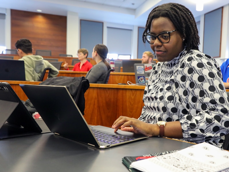 A woman wearing glasses and a patterned blouse works on a laptop in a classroom, surrounded by other students. She is focused and engaged, with a notebook and pens beside her.