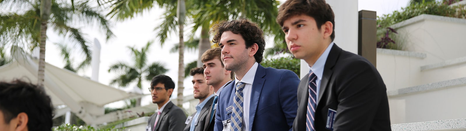 Business students in professional attire sitting outside with palm trees behind them.