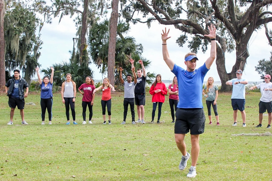 A group of students outdoors in a grassy area following a leader who has his hands raised, participating in a team-building activity surrounded by trees.