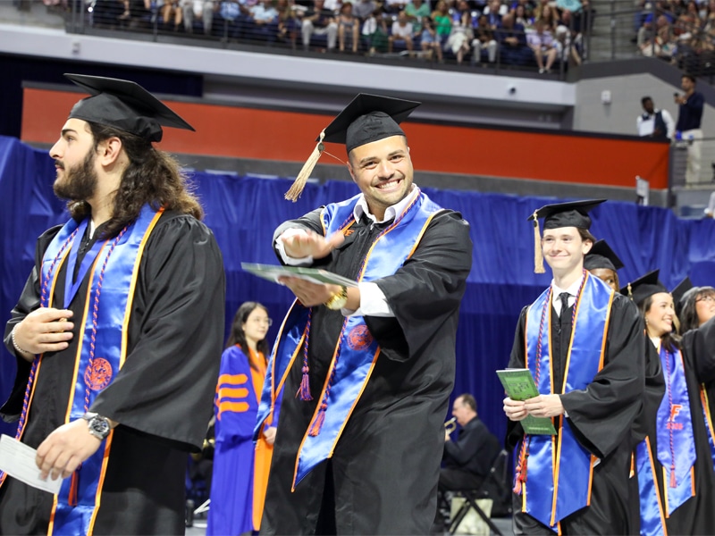 A smiling graduate in a cap and gown holding his diploma, with other graduates in line at the Heavener School of Business commencement ceremony.
