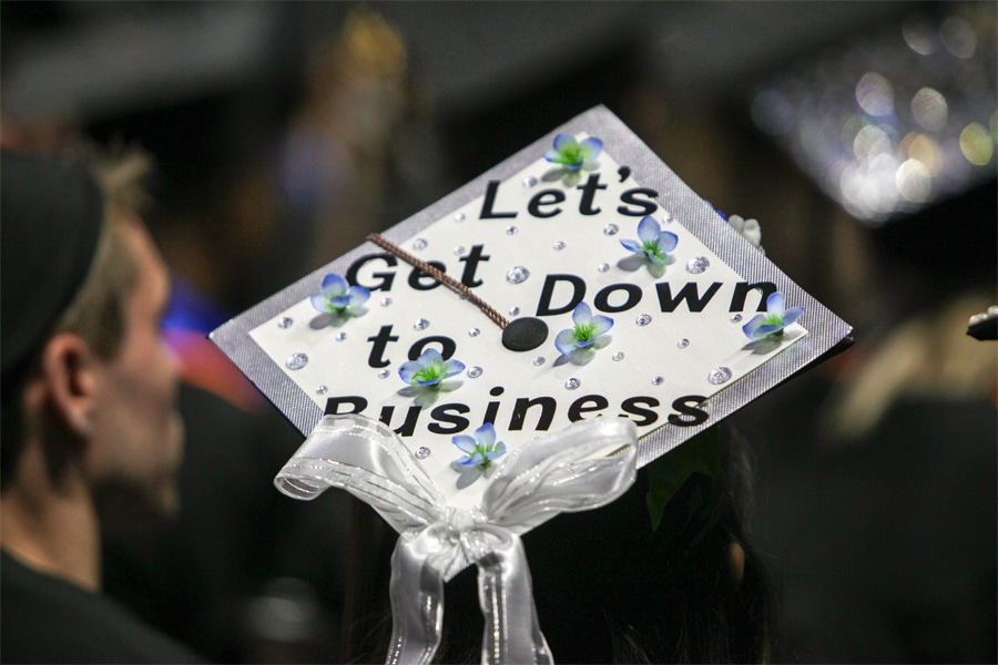 Overhead view of a decorated graduation cap reading “Let’s Get Down to Business,” adorned with small blue flowers, rhinestones, and a white ribbon bow.