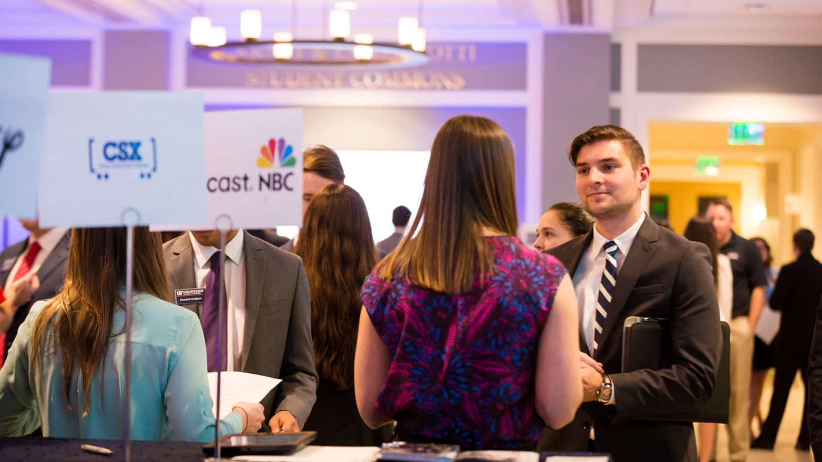 Students in business attire interact with recruiters at a career fair, with banners for companies like CSX and NBC visible.